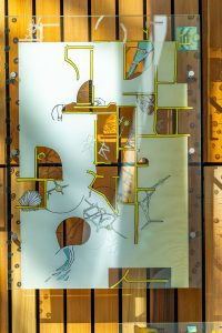 One of the glass panels of the Lines of Sight art piece. The lines on the glass cast shadows on the wood slat wall behind the panel, showing the same design and giving a three-dimensional effect.
