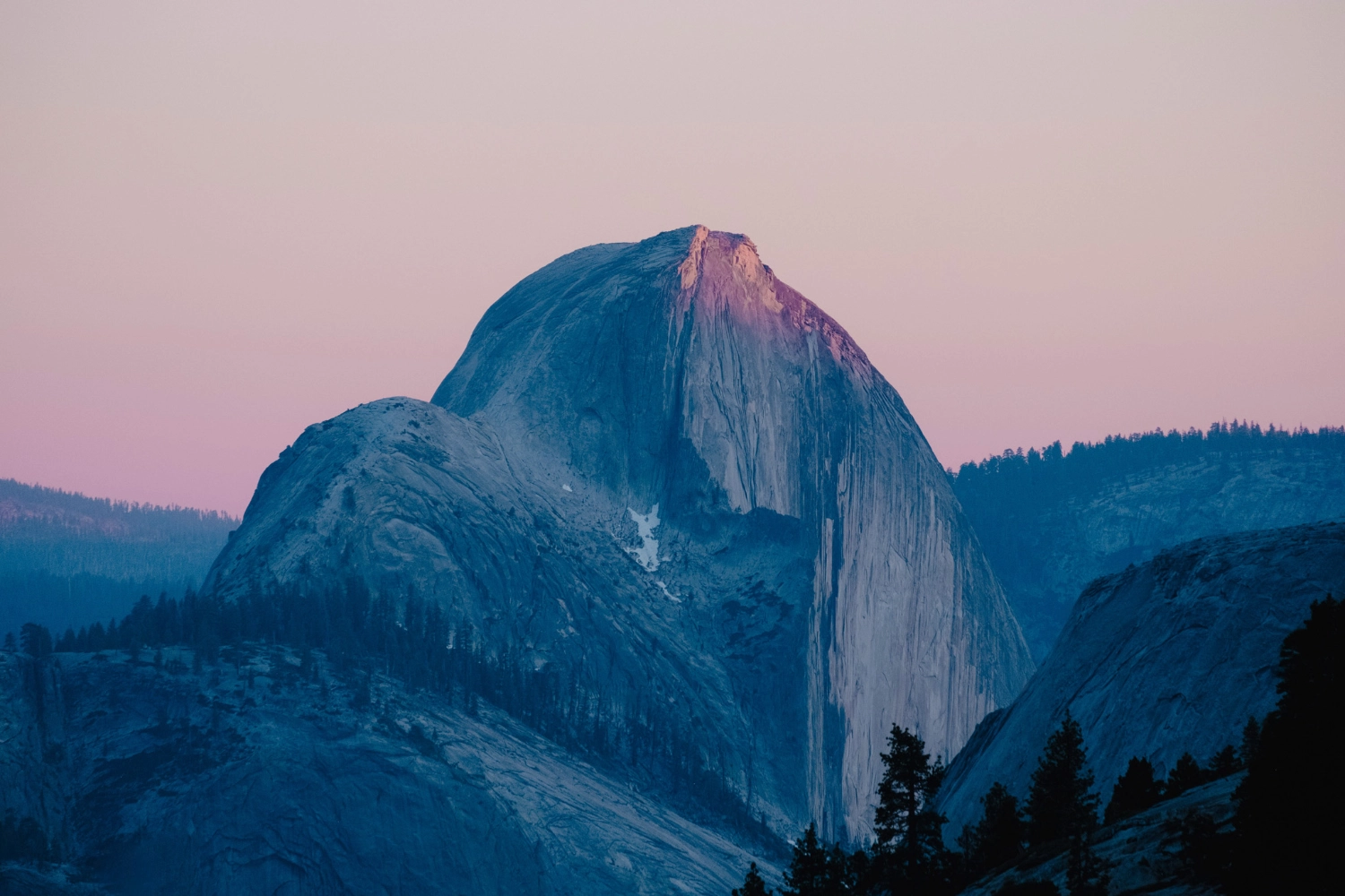The iconic Half Dome mountain in Yosemite National Park. It is illuminated by the soft pink and orange hues of a sunrise or sunset.