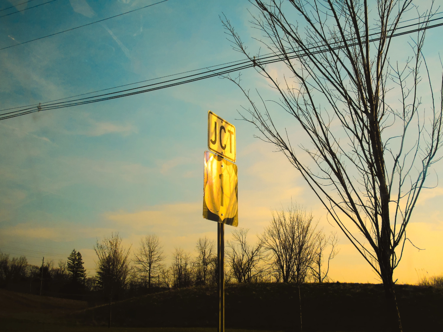 A junction sign with "JCT" and the number "1" is illuminated by the golden light of sunset. Bare trees and power lines are silhouetted against the sky.